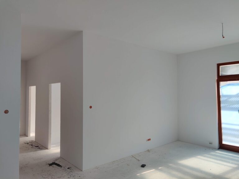 Wnętrze nowego mieszkania w stanie deweloperskim podczas odbioru technicznego; widoczne świeżo otynkowane ściany, otwory drzwiowe bez futryn oraz prowizoryczne instalacje elektryczne.